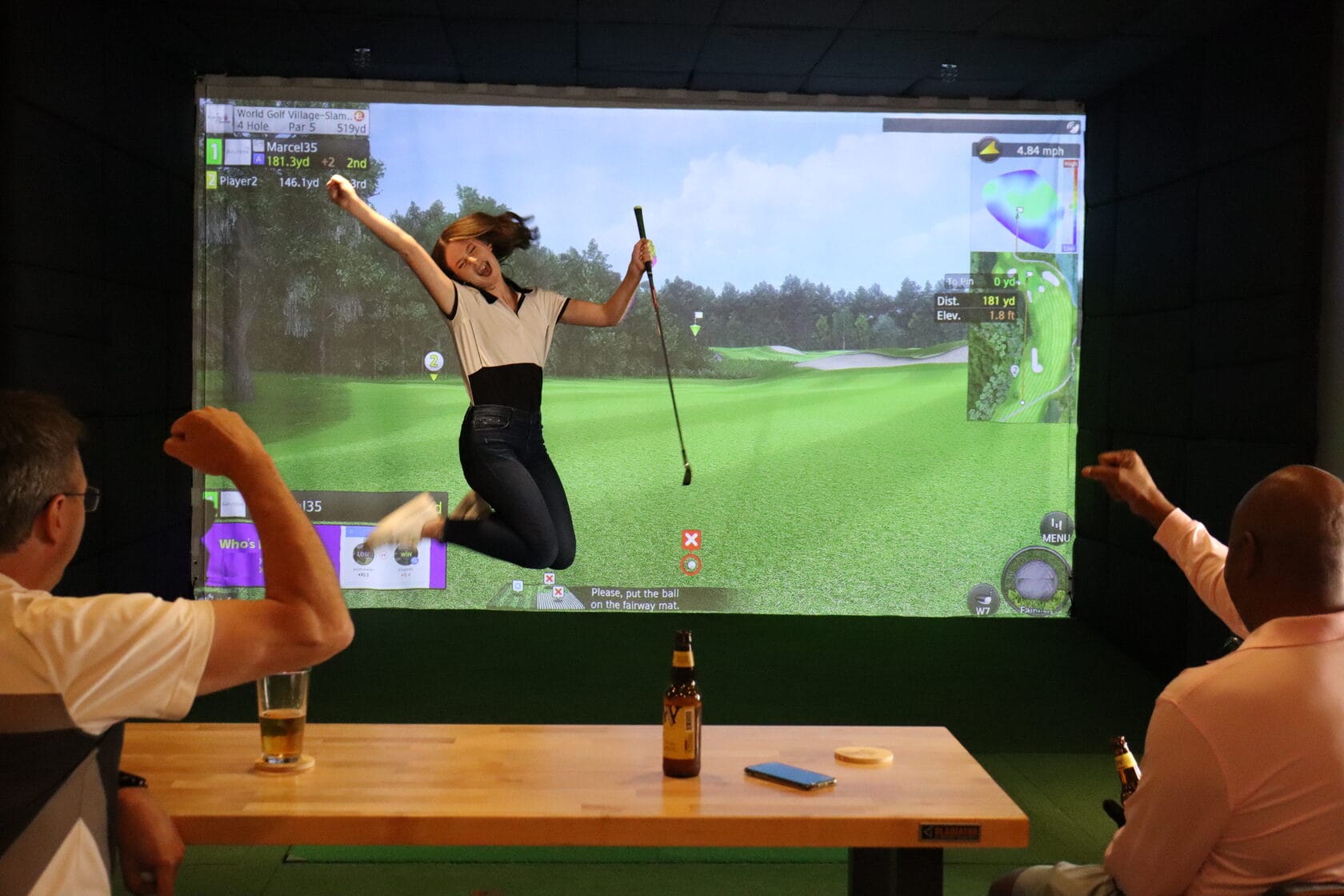 A golfer leaping in celebration in front of a simulator screen while friends at the table behind cheer